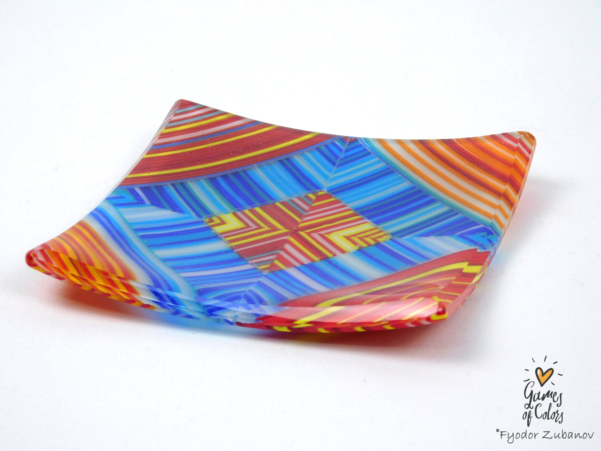 A square, fused glass plate with a vibrant multicolor geometric pattern of blue, red, orange, yellow, and white stripes, shown on a plain white background.