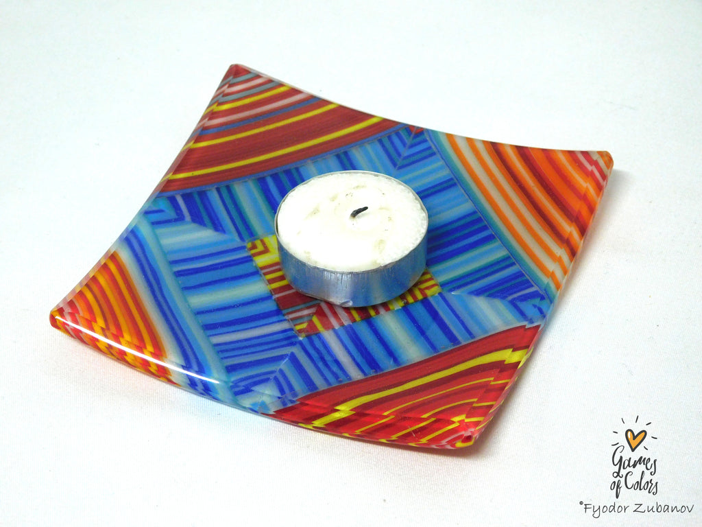 A square glass candle holder with a colorful geometric pattern of blue, red, orange, and yellow stripes, holding a small white tea light candle on a white background.