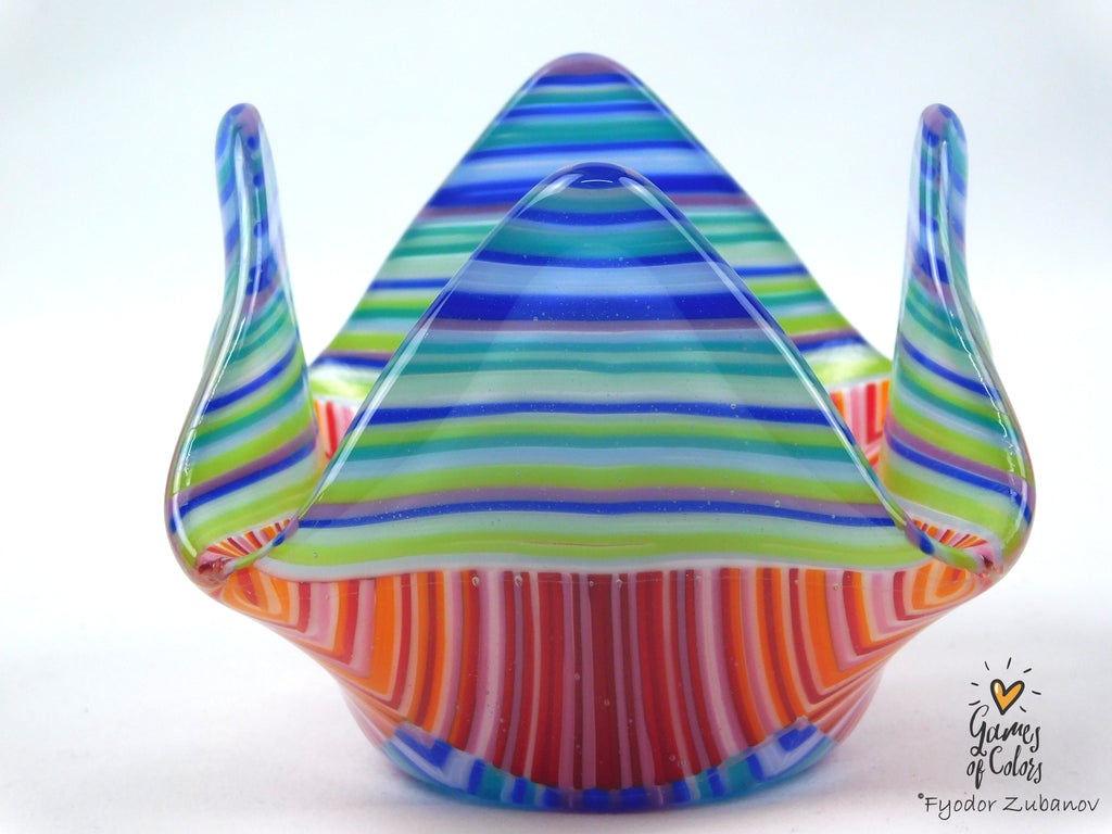 A colorful fused glass candle holder with a mountain-like flared rim, featuring horizontal striped patterns in blue, green, orange, red, and white on a white background.