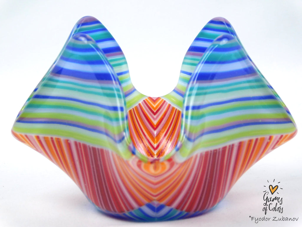 A colorful hand-blown glass sculpture with a symmetrical, wing-like or butterfly-inspired shape, featuring vibrant horizontal and vertical striped patterns in blue, green, orange, red, and white on a white background.