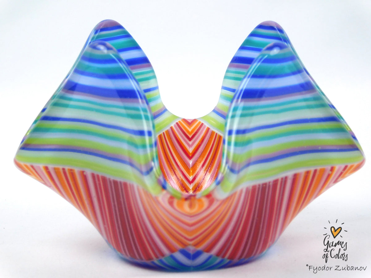 A colorful hand-blown glass sculpture with a symmetrical, wing-like or butterfly-inspired shape, featuring vibrant horizontal and vertical striped patterns in blue, green, orange, red, and white on a white background.