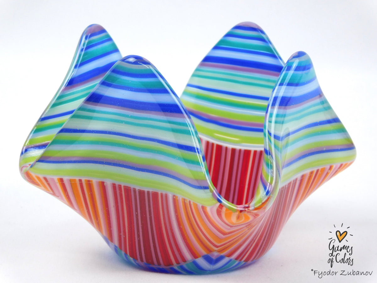 A colorful hand-blown glass bowl with wavy, mountain-like ridges, featuring horizontal blue, green, and white stripes on the upper half and vertical red, orange, and white stripes on the lower half, set against a plain white background.
