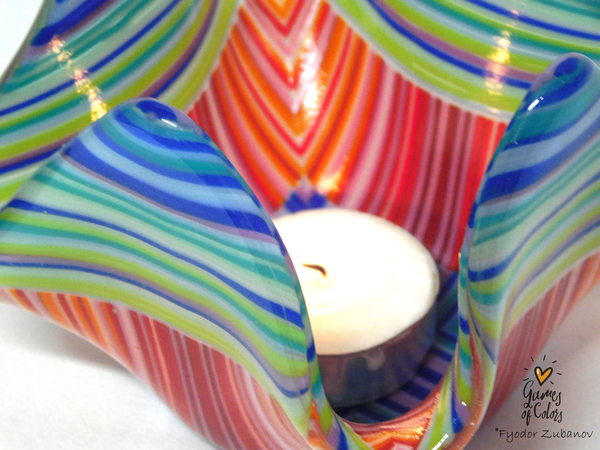 A close-up of a colorful fused glass holder with swirling stripes of blue, green, orange, pink, and white, containing a white tea light candle.