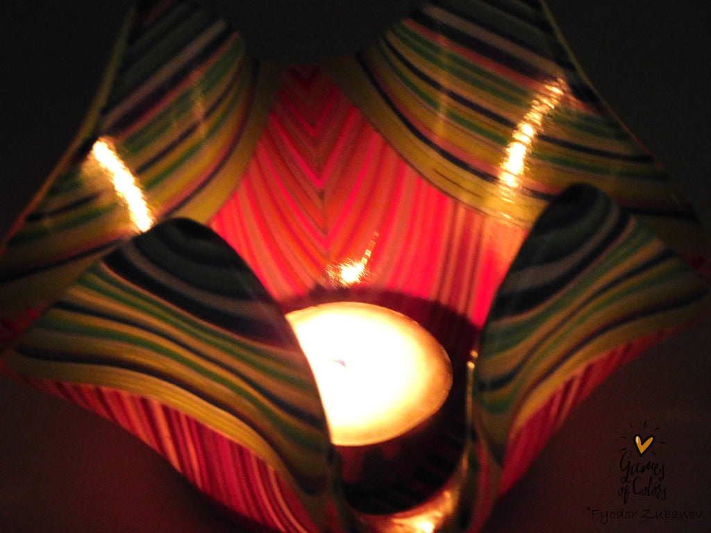 A close-up of a colorful fused glass candle holder with a lit tealight candle inside, set against a dark background.