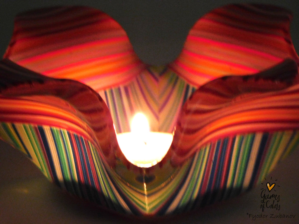 A handmade glass candle holder with a wavy, scalloped edge, decorated with colorful vertical stripes in red, orange, pink, green, blue, and purple. A lit tea light candle is placed inside the center, casting a warm glow.