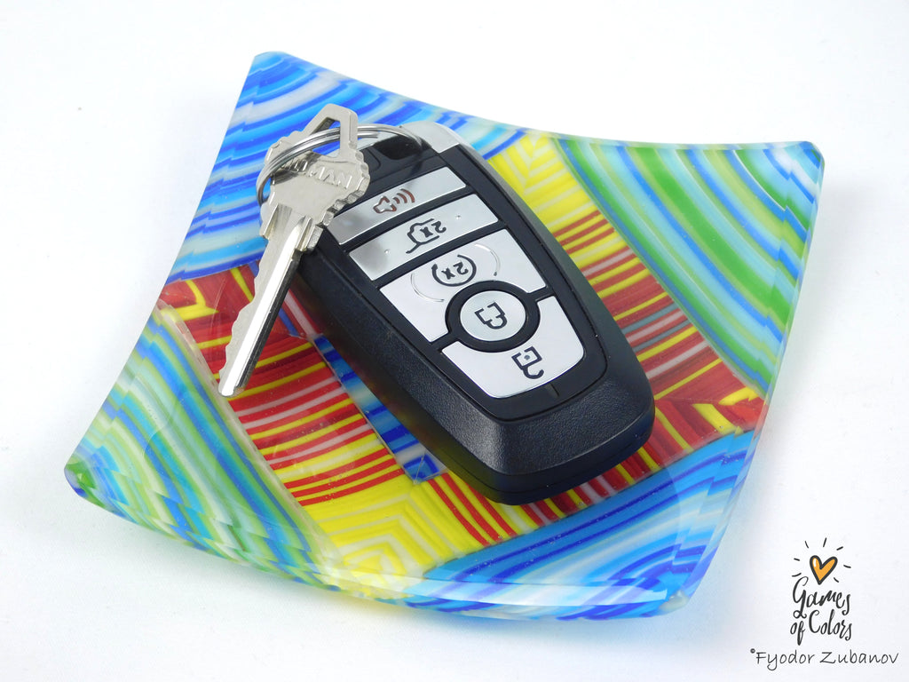 A black car key fob with keys resting on a colorful, square, abstract-patterned glass plate. The plate features swirls of blue, green, yellow, and red on a white background.