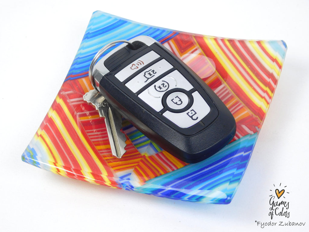 A colorful square glass dish with a vibrant multicolor swirl pattern in blue, red, yellow, and orange, holding a black car key fob and keys. The dish is photographed on a clean white background with a 'Games of Colors' watermark in the bottom right corner.