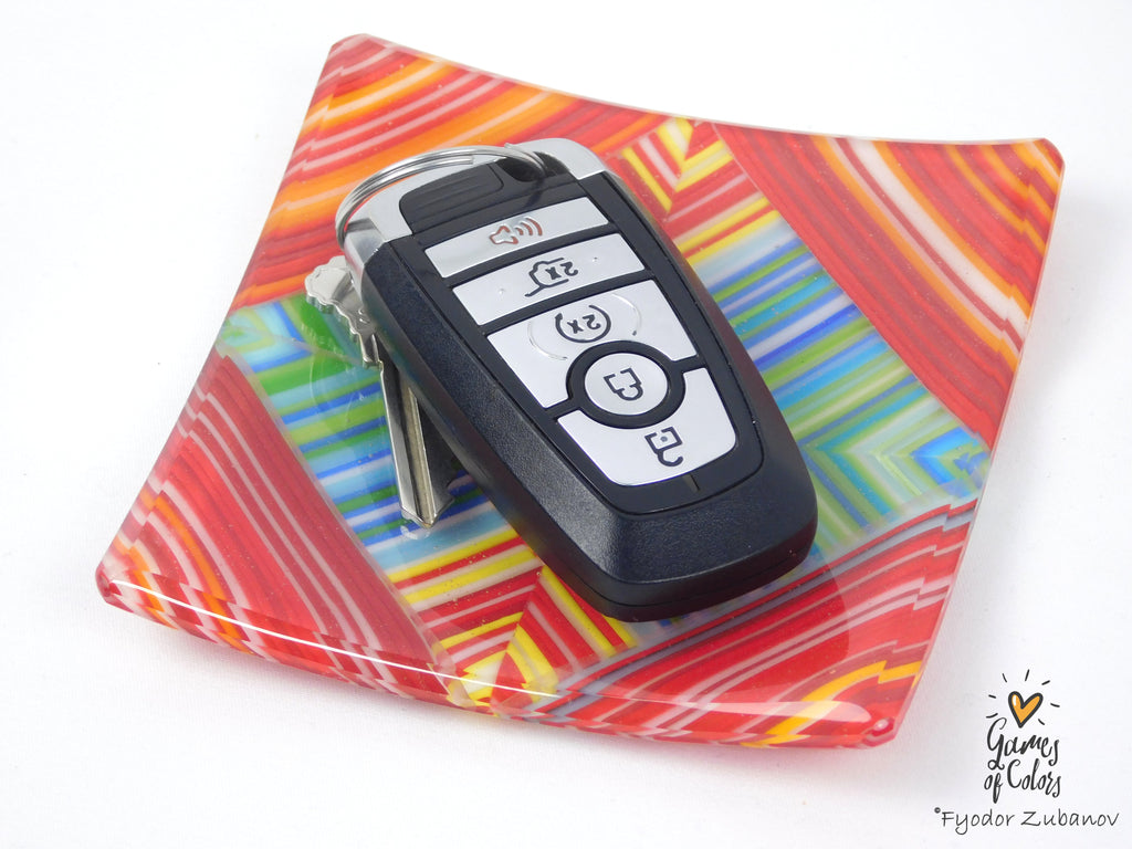 A black car key fob with silver keys resting on a square, colorful glass plate with a red, orange, yellow, and blue geometric pattern, set against a plain white background.