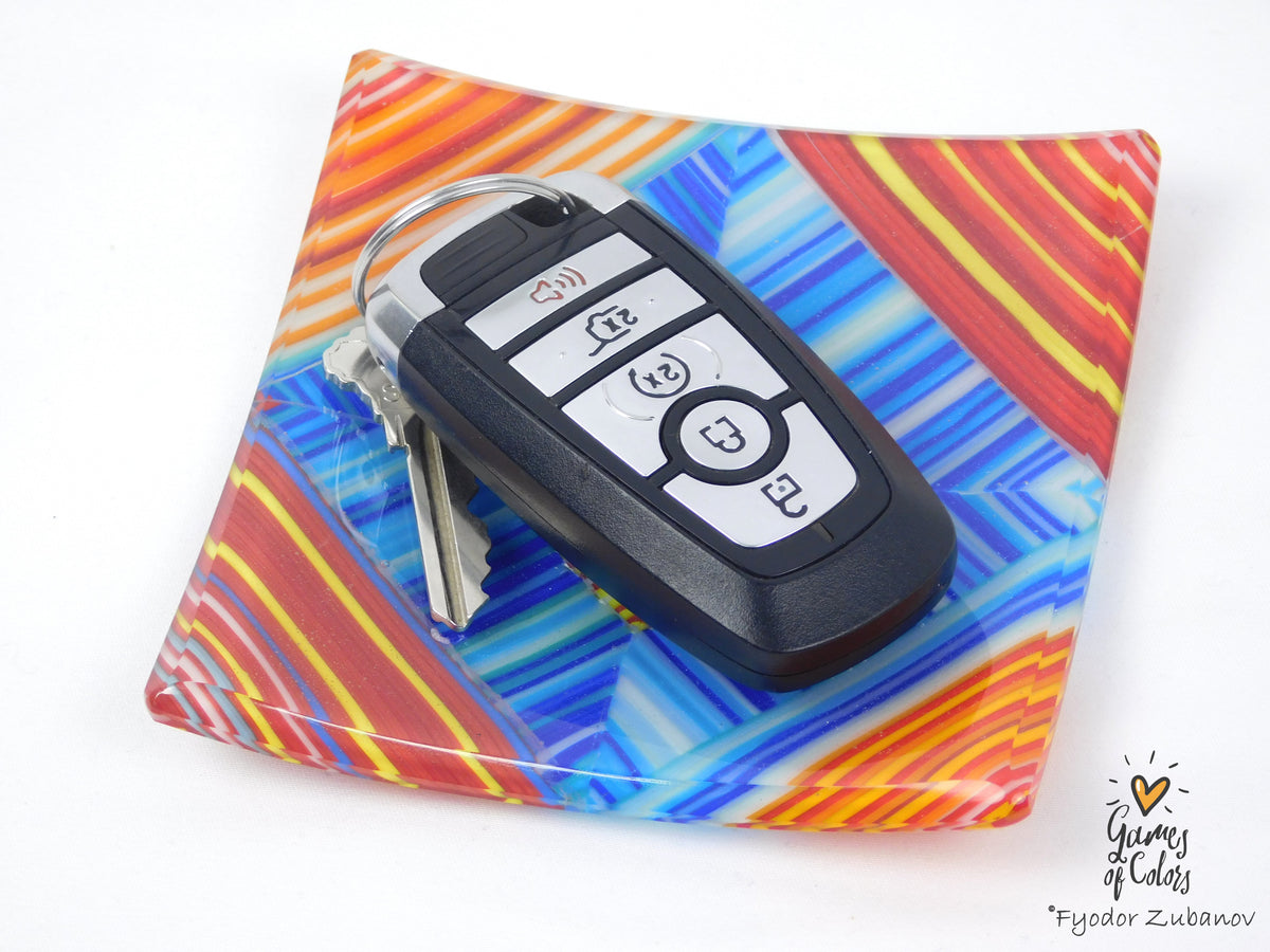 A black car key fob with keys attached, displayed on a square glass plate with a vibrant multicolor abstract pattern of red, orange, yellow, blue, and white on a white background.