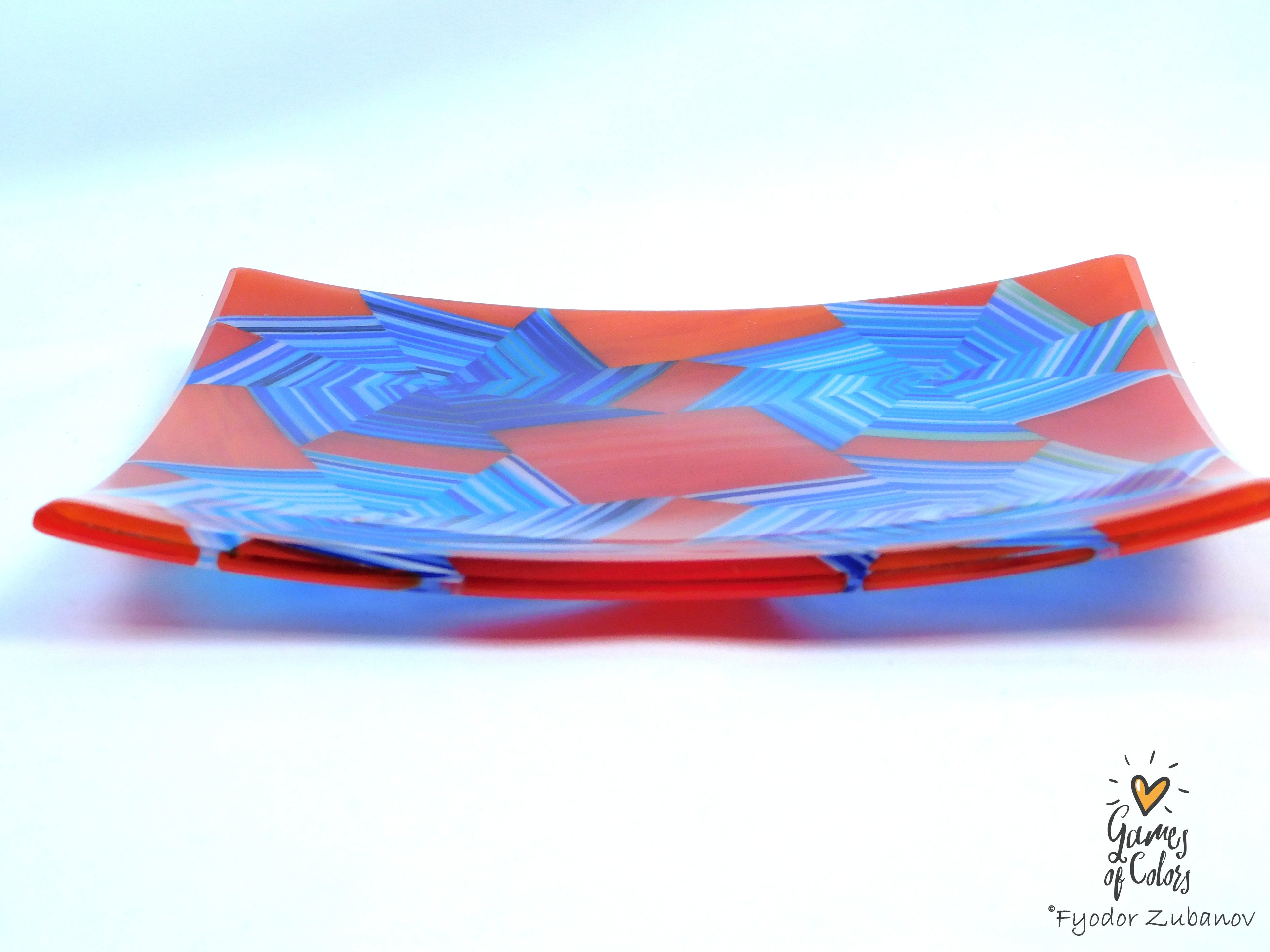 A square glass serving platter with a vibrant red-orange base and a geometric blue starburst pattern, photographed on a clean white background with a soft shadow.