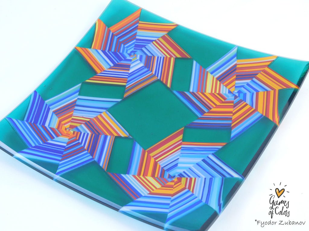 A square glass plate featuring a colorful geometric starburst pattern with concentric stripes in shades of blue, orange, red, and yellow on a teal green background, photographed on a white surface. Branded Fyodor Zubanov, Games of Colors