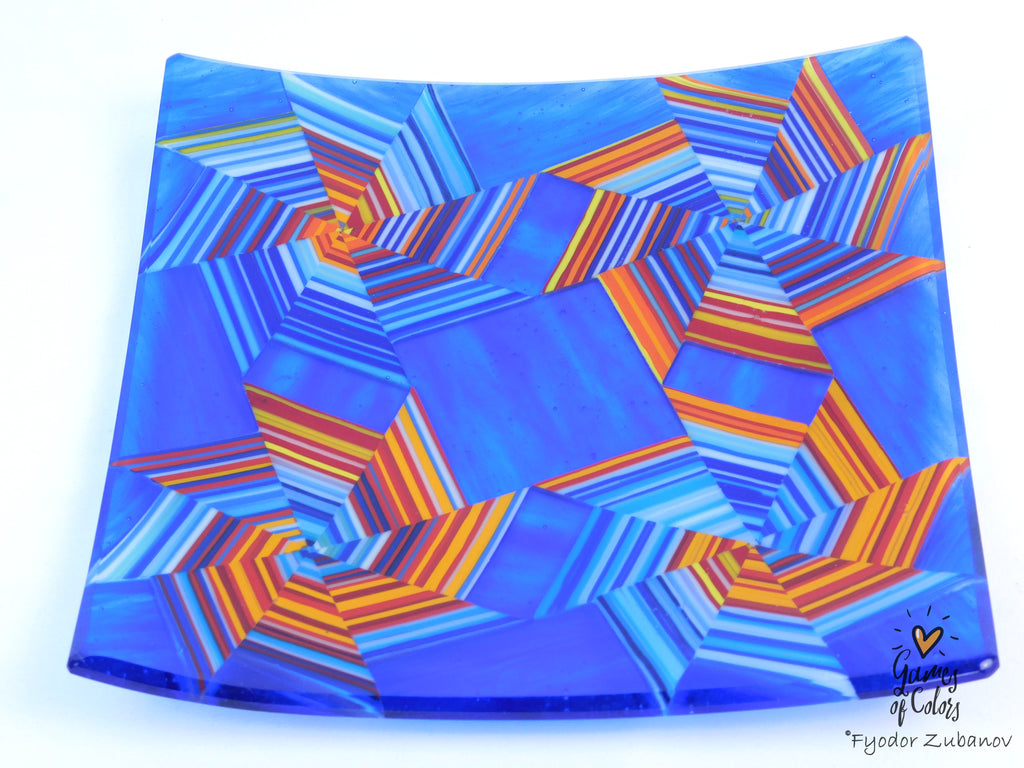 A square glass plate featuring a vibrant geometric pattern of 3D-effect segments in shades of blue, orange, and red, photographed on a clean white background. Featured Fyodor Zubanov, Games of Colors