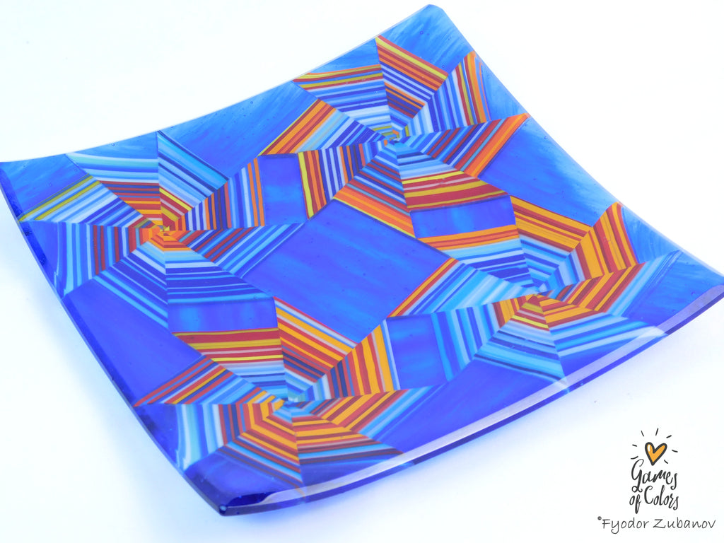 A square glass plate featuring a vibrant blue and orange geometric starburst pattern, photographed on a clean white background with a 'Games of Colors' watermark in the bottom right corner.