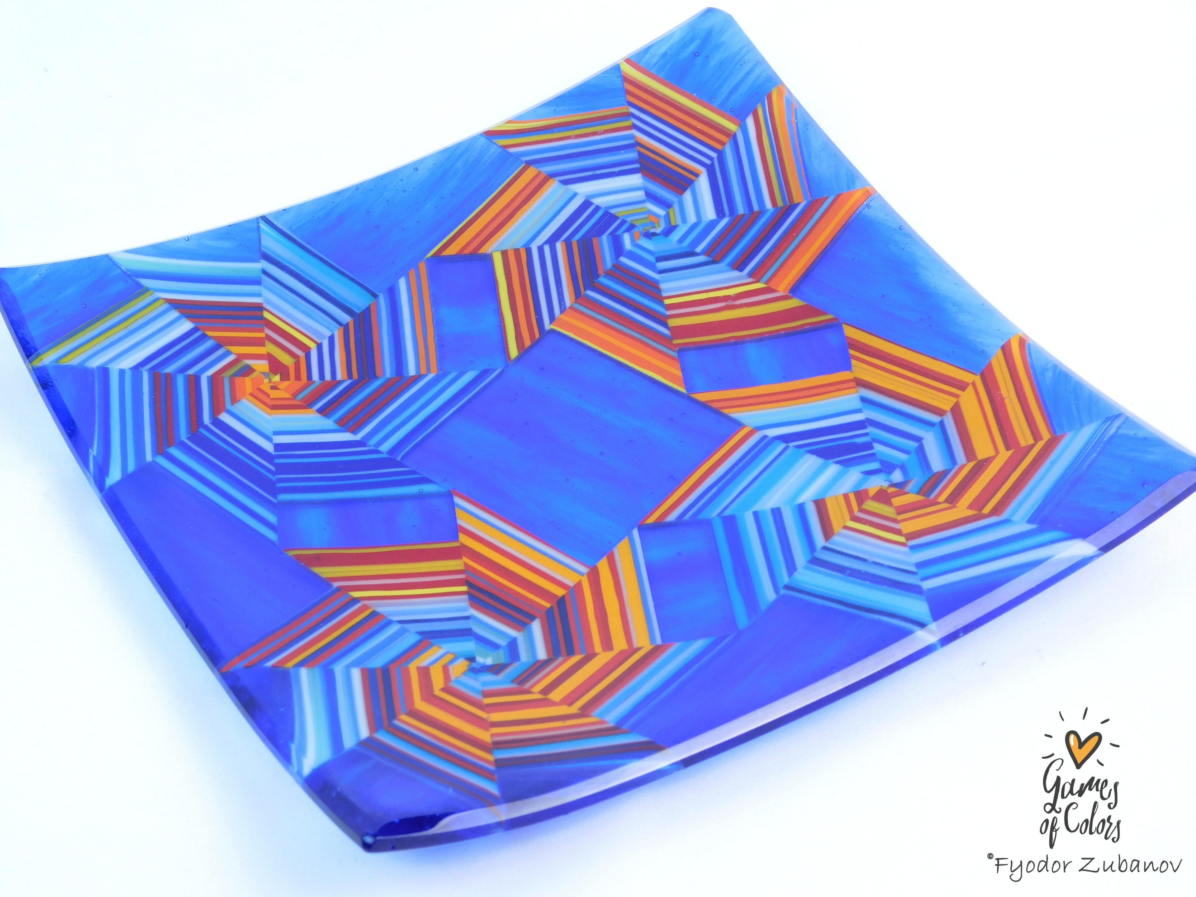 A square glass plate featuring a vibrant blue and orange geometric starburst pattern, photographed on a clean white background with a 'Games of Colors' watermark in the bottom right corner.