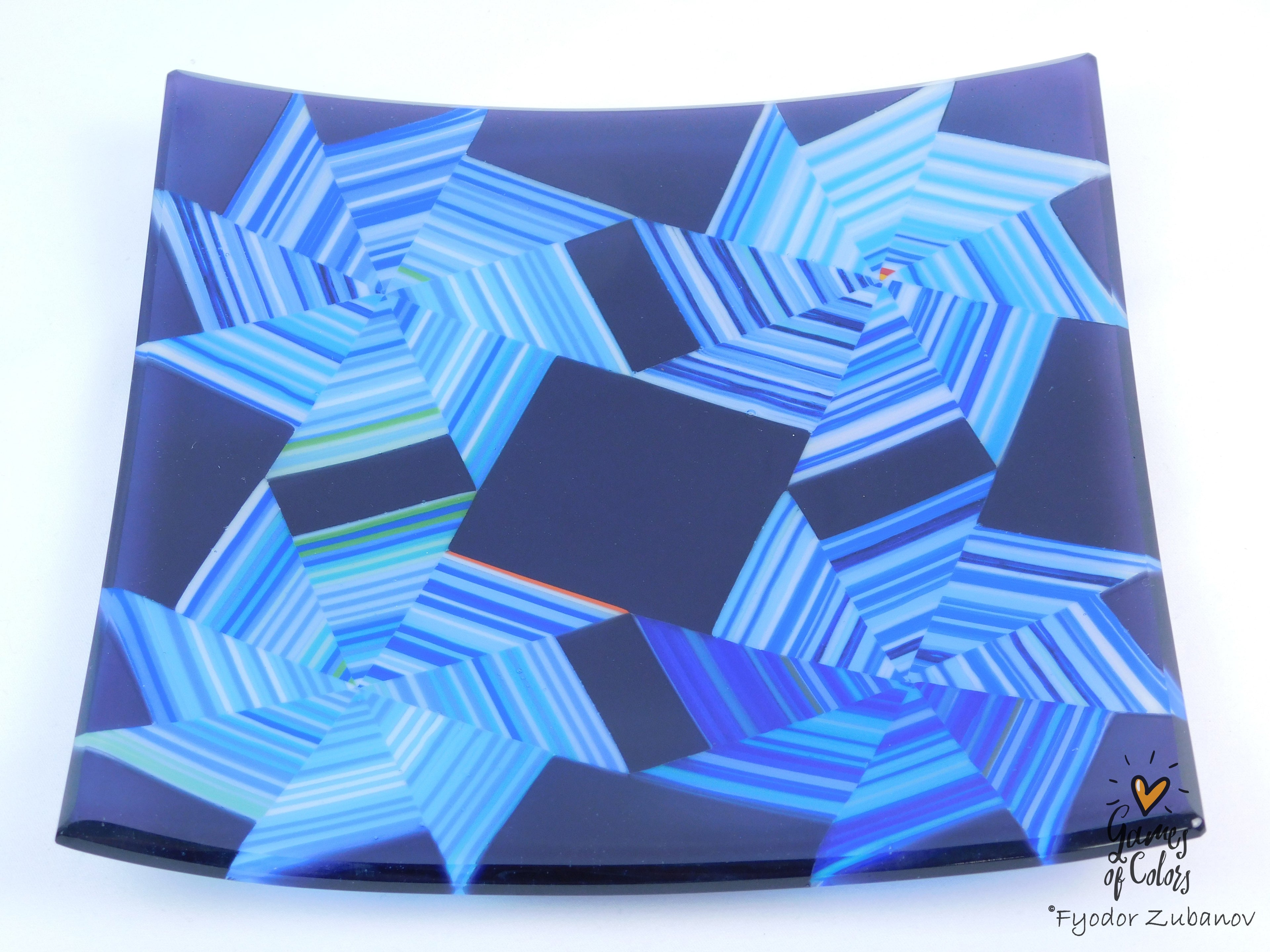 A square glass plate featuring an intricate geometric starburst pattern in shades of blue, navy, and light blue, with a small artist signature in the bottom right corner.