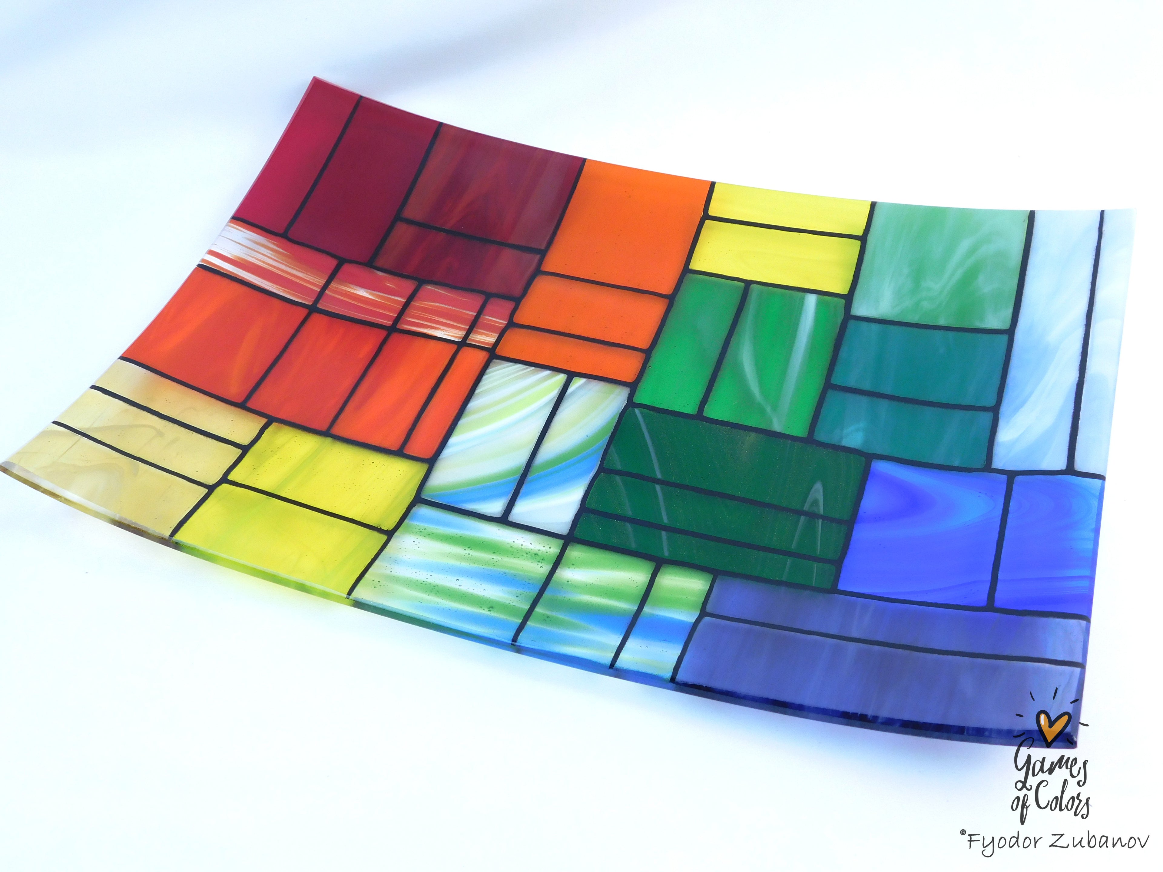 A colorful stained-glass-style square plate with geometric color blocks outlined in black, photographed on a white background. The plate contains multiple colors including red, orange, yellow, green, blue, and purple, with some marbled swirls.