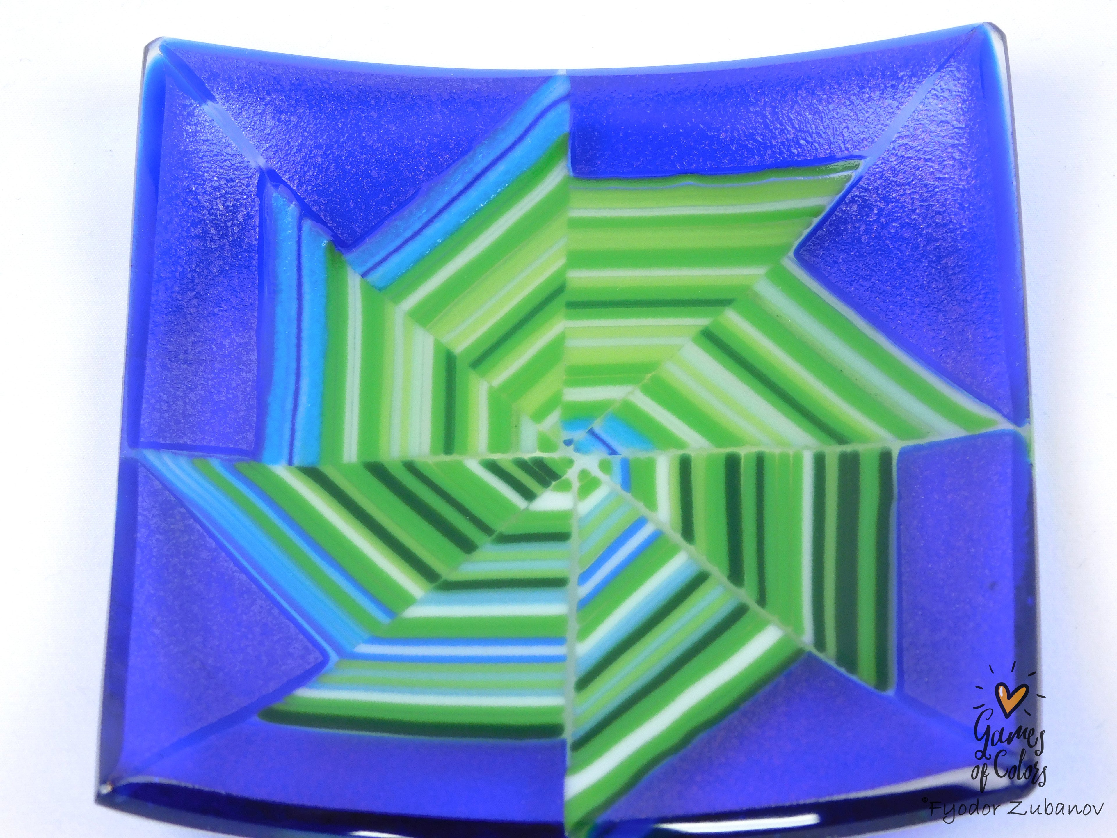 A square glass plate with a geometric starburst pattern in vibrant blue and green, featuring a glossy finish on a white background.