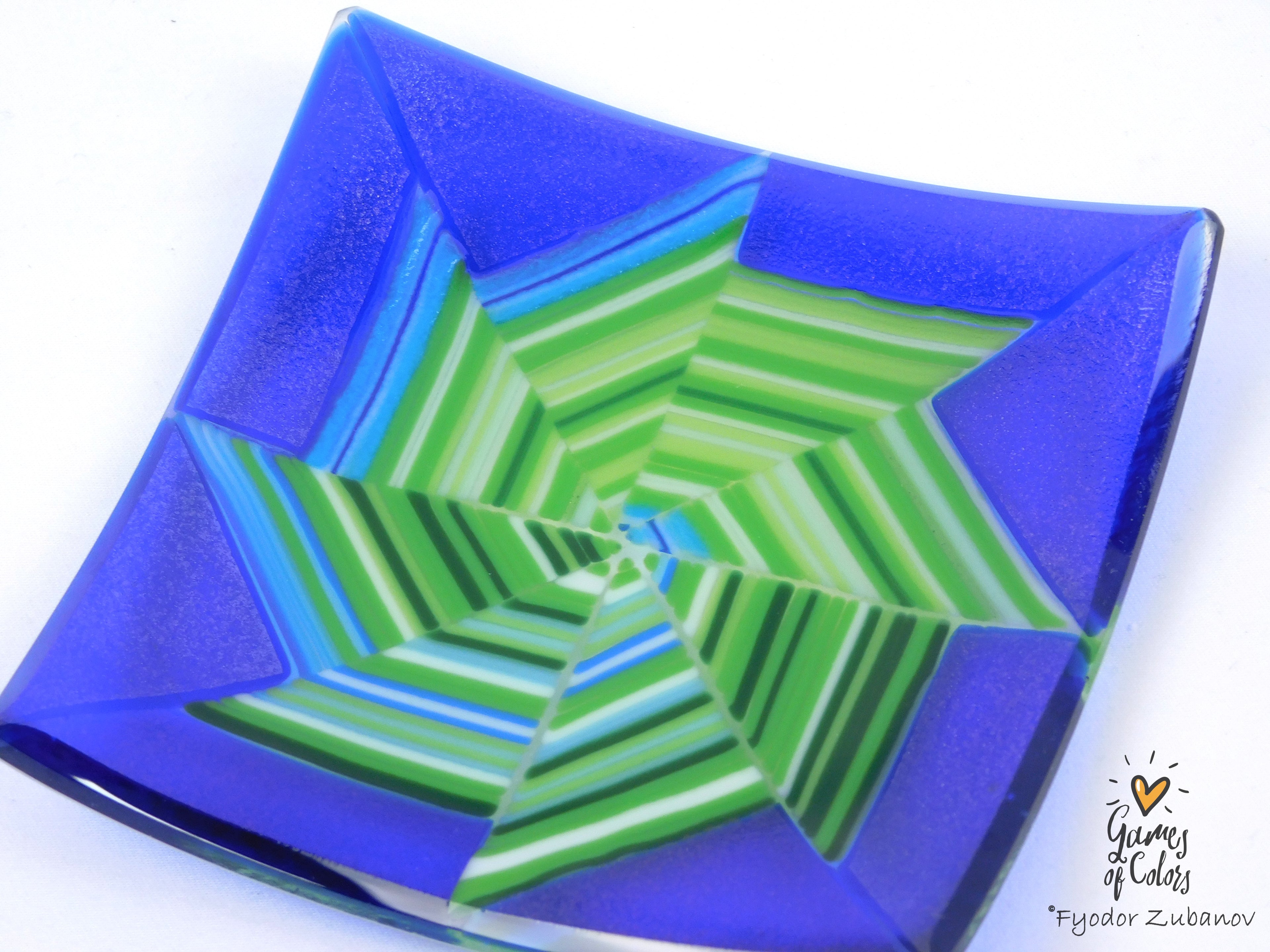 A square fused glass plate featuring a geometric starburst pattern with concentric rings of green, blue, and white stripes on a vibrant cobalt blue background, shown on a white background.