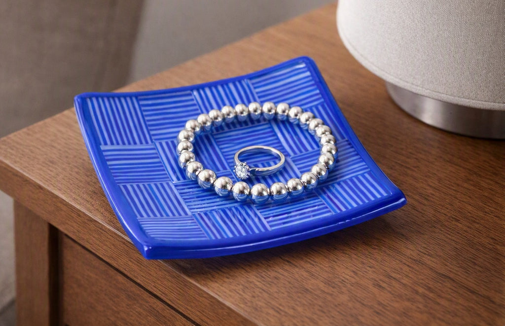 A silver beaded bracelet with a small clear stone ring resting on a textured blue square dish, placed on a wooden table next to a lamp.