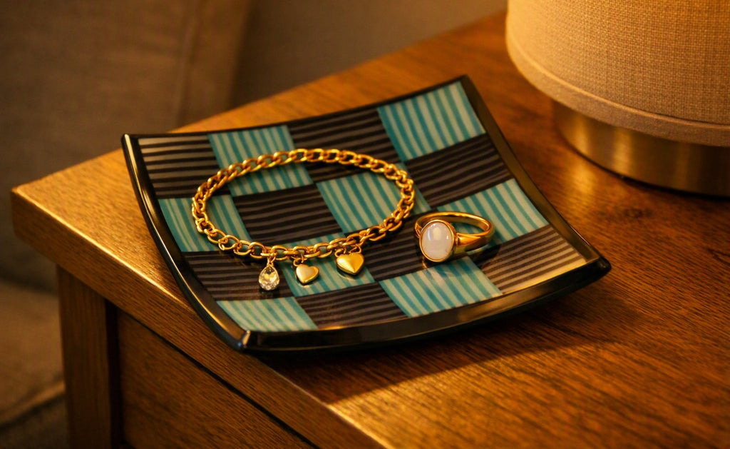 A gold chain bracelet with heart charms and a gold signet ring with a white stone, displayed on a black and teal striped tray resting on a wooden table next to a lamp.