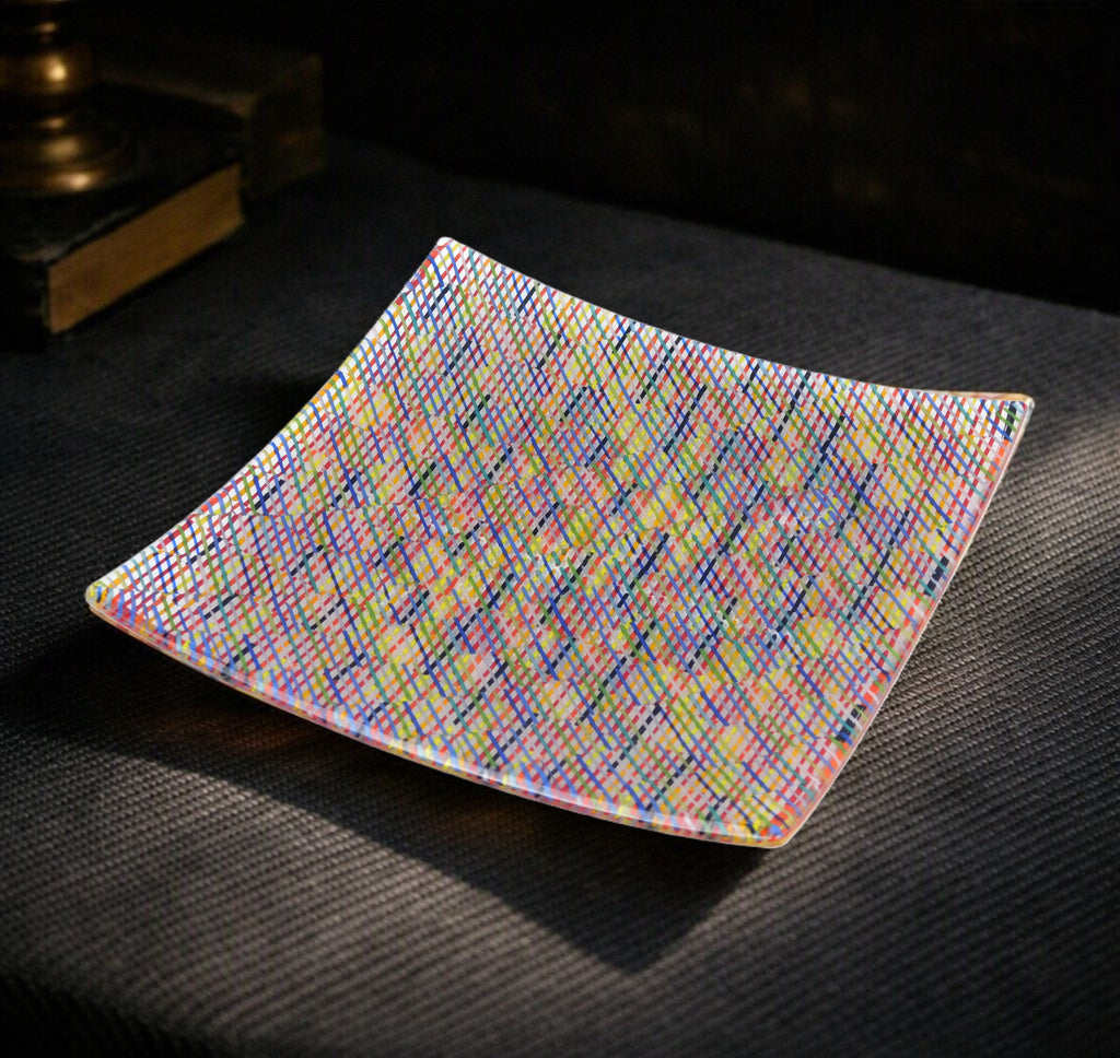 A square glass plate with rounded corners, featuring a vibrant, multicolored rainbow geometric pattern, resting on a dark textured surface.