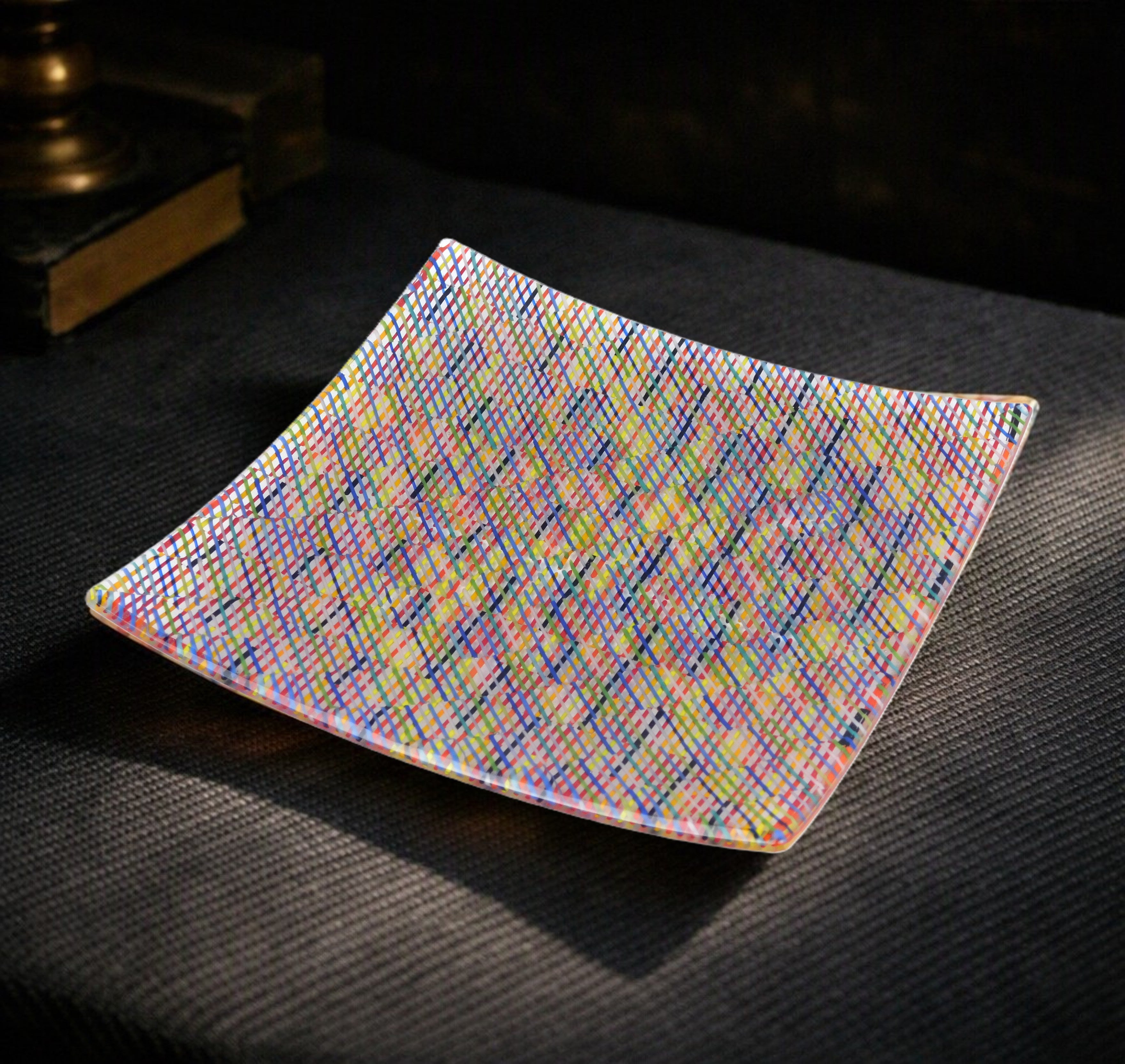 A square glass plate with rounded corners, featuring a vibrant, multicolored rainbow geometric pattern, resting on a dark textured surface.